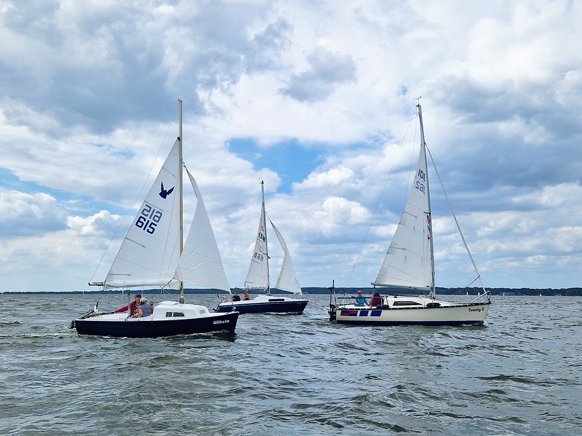 Drei Segelboote des Segel-Klub Minden segeln nebeneinander auf einem See unter leicht bewölktem Himmel. Die Boote sind in voller Fahrt, und auf jedem Boot sind mehrere Segler zu sehen. Das Wasser ist leicht wellig, und die Atmosphäre wirkt entspannt und lebendig.