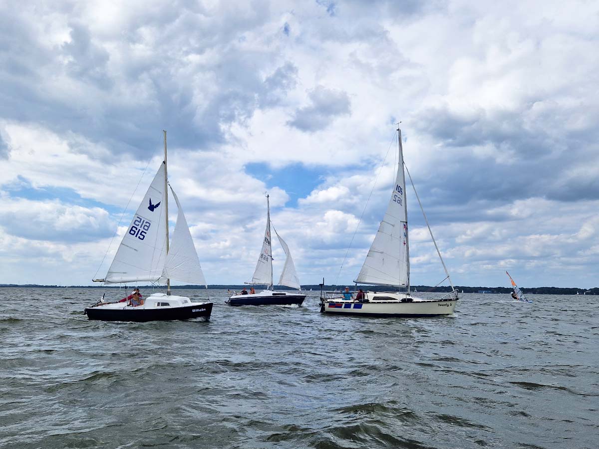 Drei Segelboote des Segel-Klub Minden segeln nebeneinander auf einem See unter leicht bewölktem Himmel. Die Boote sind in voller Fahrt, und auf jedem Boot sind mehrere Segler zu sehen. Das Wasser ist leicht wellig, und die Atmosphäre wirkt entspannt und lebendig