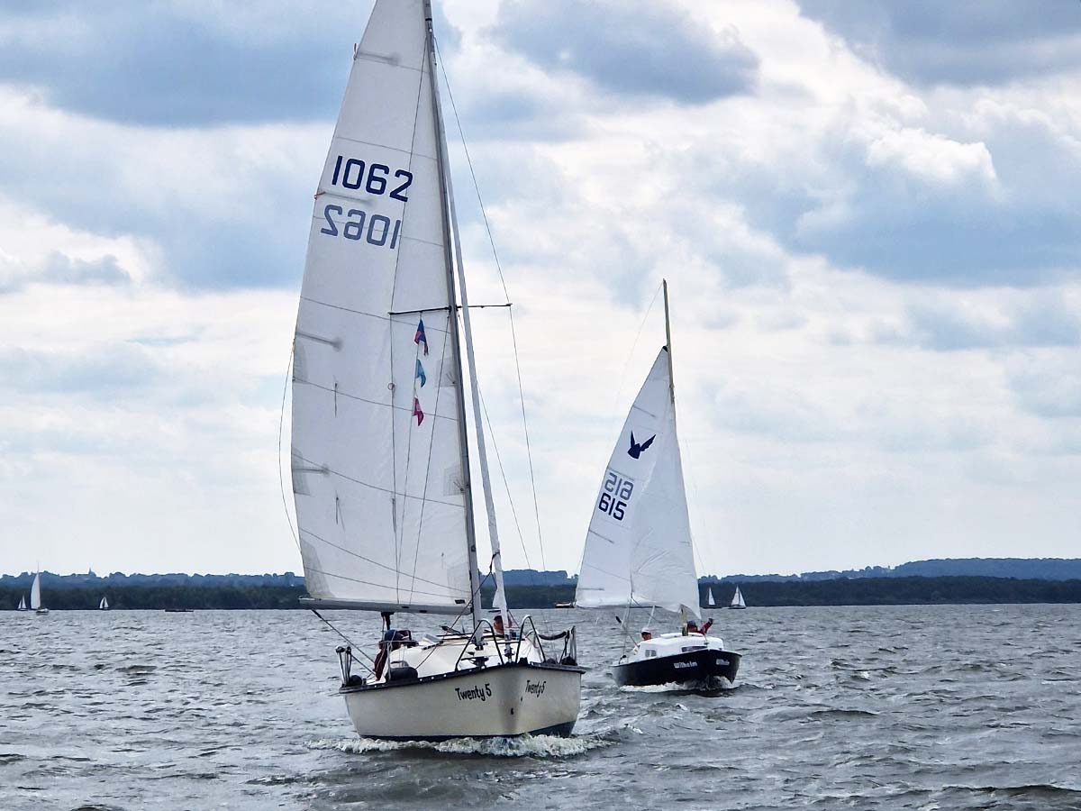 Zwei Segelboote segeln bei leicht bewölktem Himmel auf einem See. Im Vordergrund ist das Boot 